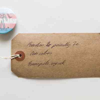 A badge which says 'Out And Proud' with a trans flag as the background and a figure.