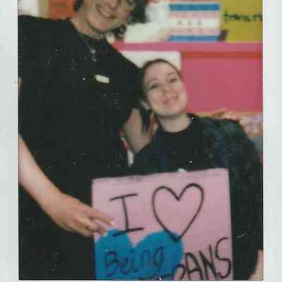 Two people smiling happily hold a pink sign that reads, "I love being trans," against a colorful background.