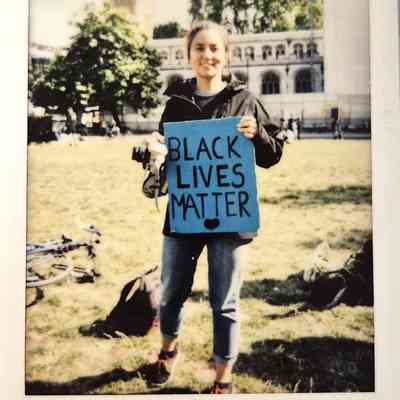 A person is holding a "Black Lives Matter" sign outdoors near a historic building and trees.