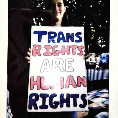 A person holds a sign reading "TRANS RIGHTS ARE HUMAN RIGHTS," with trees in the background.