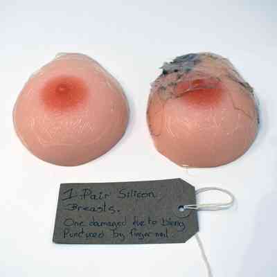 The image shows a pair of silicone breasts, one damaged, with a handwritten tag describing them.