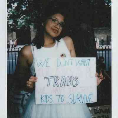A person holds a sign which says "We Don't Want Your Cis Kids To Be Trans We Want Your Trans Kids To Survive" standing outdoors, near trees, wearing glasses and a white dress.