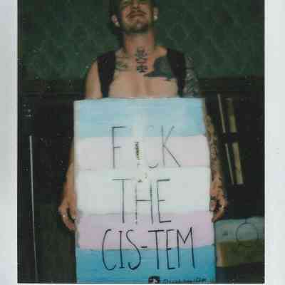 A person with tattoos is holding a sign with text in a dimly lit room.