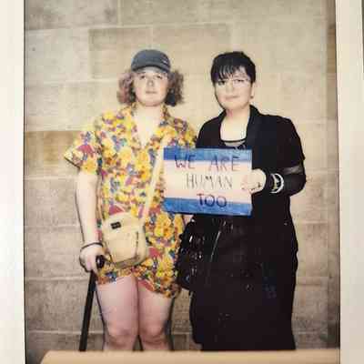 Two people stand together, one holding a sign saying “We Are Human Too.”