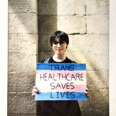 A person holding a colorful sign reads, "Trans healthcare saves lives," against a brick wall background.