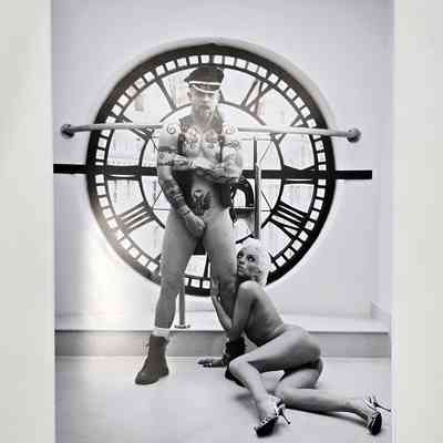 Black and white photograph of two people. One is stood up and only wearing a waistcoat and a hat, the other is on the floor leaning against the other and is naked.