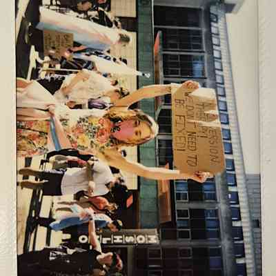 A person in a floral dress and mask holds a protest sign which says "BAN CONVERSION THERAPY WE DON'T NEED TO BE FIXED".