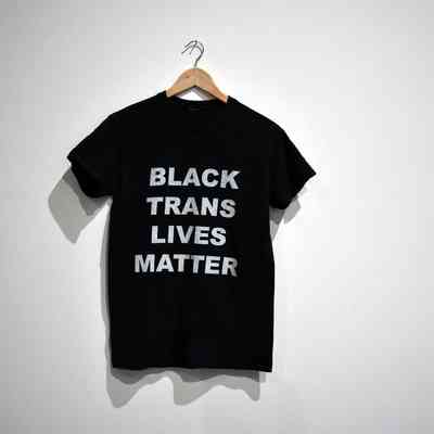 A black T-shirt hangs on a wooden hanger, with the text “Black Trans Lives Matter” in bold white text.