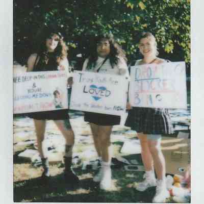 Three people stand together outdoors, holding signs supporting trans youth, under a tree with sunlight filtering.