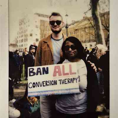 Two individuals at a public event hold a "Ban All Conversion Therapy" sign while standing closely together.