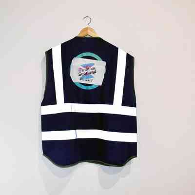 A dark vest with white reflective stripes, which says 'Trans Pride Scotland Steward' hangs from a wooden hanger on a plain wall.