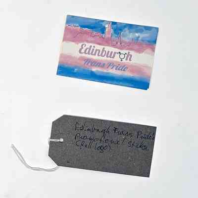 Rectangular sticker with the text "Edinburgh Trans Pride" (the 'g' in Edinburgh has been replaced with the trans symbol). The background is in the trans colours with a line drawing of a city skyline. With a brown, handwritten luggage tag.