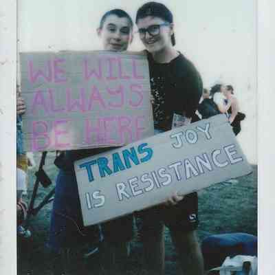 Two individuals holding signs read, "We will always be here" and "Trans joy is resistance."