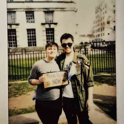 Two people stand together smiling, holding a sign which says "TRANS RIGHTS NOW".