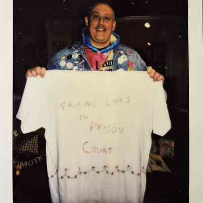 A person holding a shirt with the message "TRANS LIVES IN PRISON COUNT," smiling brightly.