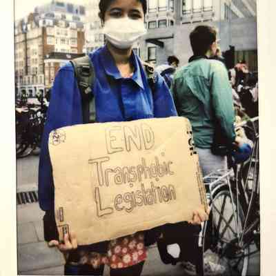 A person wearing a mask holds a sign that reads, "END Transphobic Legislation".