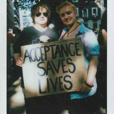 Two people are holding a cardboard sign that says "Acceptance Saves Lives" in a sunny outdoor setting.