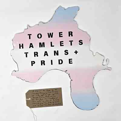 A foam board Tower Hamlets Trans+ Pride logo - which is the shape of the boundary of the London Borough of Tower Hamlets. It is colored in the blue, white and pink trans pride colours. With a brown, handwritten luggage tag.