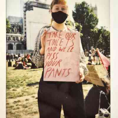 A person wearing a mask holds a sign in a park, protesting about toilet access rights for trans people.