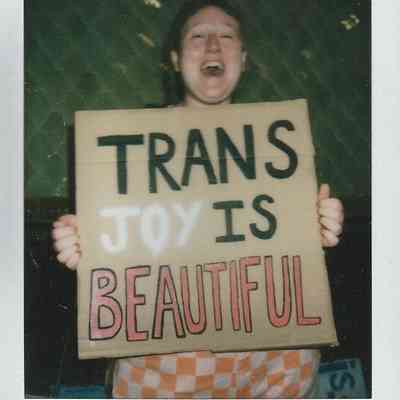 A person holds a sign reading "TRANS JOY IS BEAUTIFUL" while smiling joyfully.