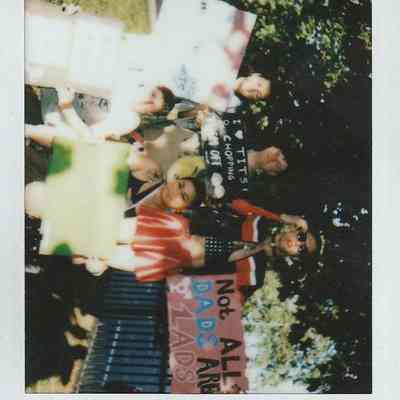 A group of people hold colorful signs outdoors, with trees visible in the background.