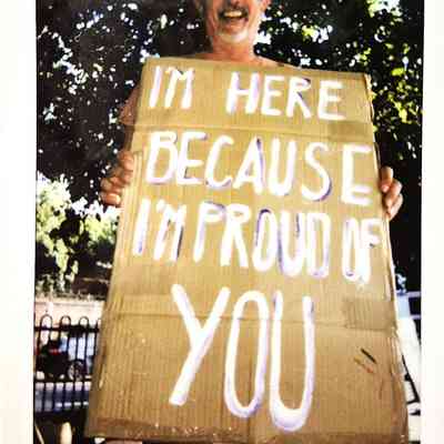 A smiling person holds a cardboard sign that says, "I'm here because I'm proud of you."