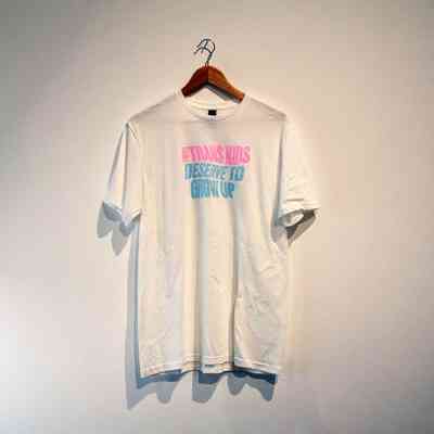 A white T-shirt with colorful text hangs on a wall emphasizing a supportive message.