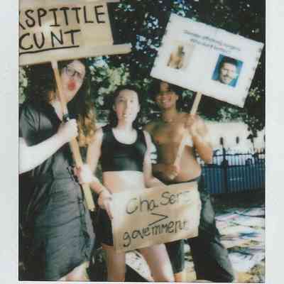 Three people hold protest signs, expressing opinions on gender and government, outdoors in a sunlit setting.