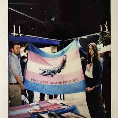 Two individuals are holding a colorful flag with text, standing at an event with tables covered in materials.