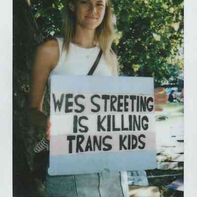 A person is holding a protest sign with bold text, standing outdoors near trees in bright daylight.