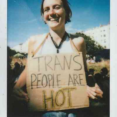A smiling person outdoors holds a sign reading "TRANS PEOPLE ARE HOT" against a sunny blue sky.