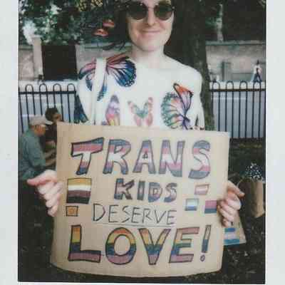 A person wearing sunglasses and a floral crown holds a colorful sign saying, "Trans kids deserve love!"