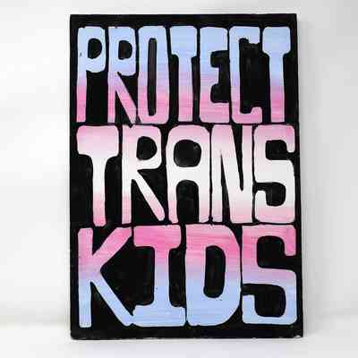 A placard which is painted black then has 'Protect Trans Kids' in large bold font in trans flag colours.