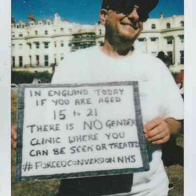 A person smiling outdoors holds a sign which says "In England today if you are aged 15 to 21 there is NO gender clinic where you can be seen or treated #FORCEDCONVERSIONNHS".