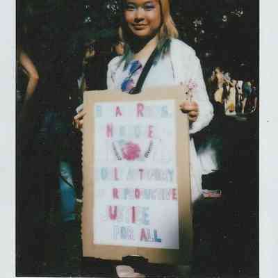 A person holds a sign which has writing on it in pink and blue but is too blurry to make out.