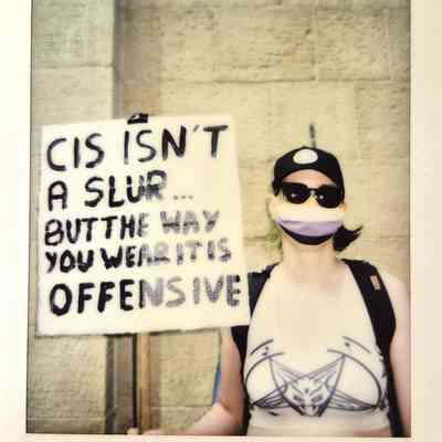 A person wearing sunglasses and a mask holds a sign reading, "Cis isn't a slur but the way you wear it is offensive".