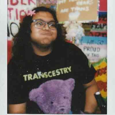 A person wearing a "TRANSCESTRY" shirt poses playfully, surrounded by colorful signs.