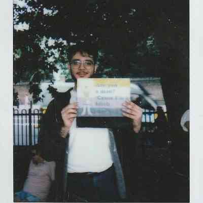 A person holding a humorous sign stands in front of a tree in a park setting.