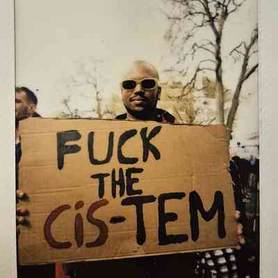A person wearing sunglasses holds a protest sign that reads, “FUCK THE CIS-TEM".