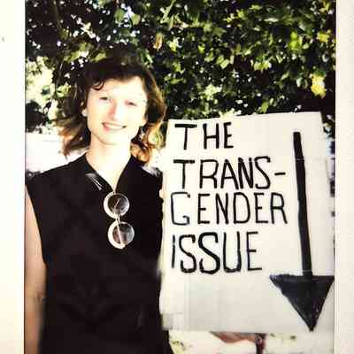 A smiling person stands beside a sign reading "The Transgender Issue" under a leafy tree canopy.