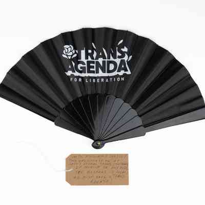 The image shows a black fan labeled "Trans Agenda for Liberation" with a handwritten tag.