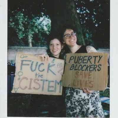 In the image, two individuals smilingly hold signs advocating for trans rights under a large tree outdoors.