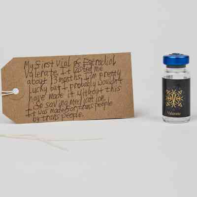Brown luggage tag with vial of Estradiol Valerate, with a blue lid, on white background.