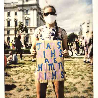 A person with sunglasses and mask holds sign for trans rights in a busy London square.