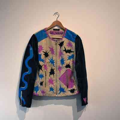 Front of leather jacket. The vest portion is white with purple sleeves. There are several paintings across the jacket - on the white front are splashes of colour. On the right arm is a blue painted snake, on the left arm a trans pride flag and pink hearts.