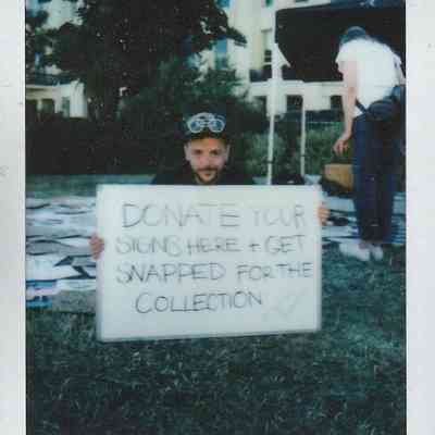 E-J Scott holds a placard which says "DONATE YOUR SIGNS HERE + GET SNAPPED FOR THE COLLECTION". Behind E-J is a grass field full of placard on the floor.