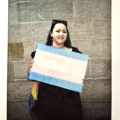 A person holds a sign with the transgender flag colors, standing against a stone wall.