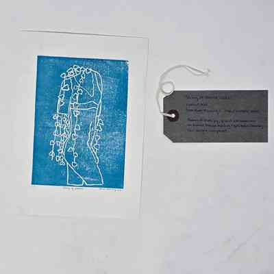 This image shows a blue linocut print depicting a headless figure with heart-shaped vines draping down from their neck. With a brow, handwritten luggage tag.