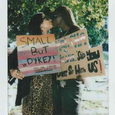 Two individuals are sharing a kiss while holding colorful protest signs under a tree.
