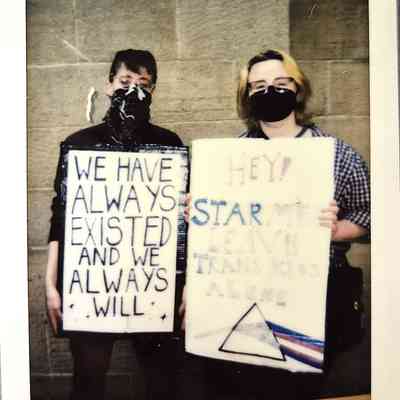 Two masked individuals hold protest signs advocating for trans rights, standing against a brick wall backdrop.